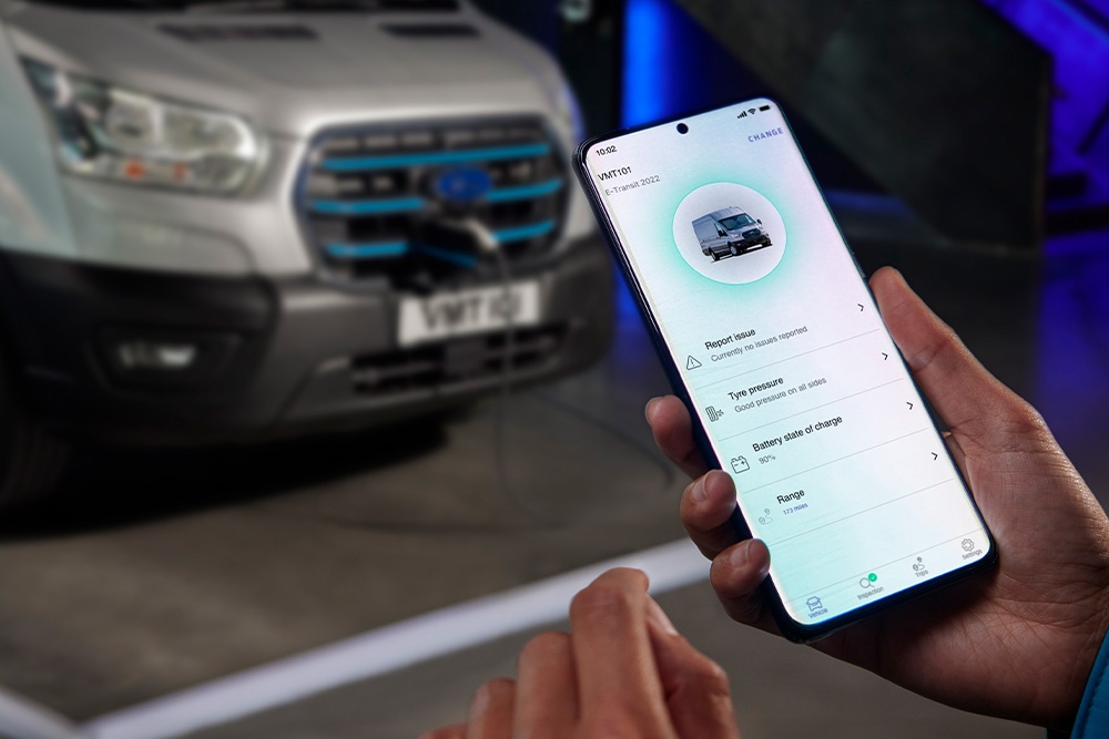 Osoba drží smartphone s aplikací Ford, v pozadí se nabíjí Ford E-Transit Custom.