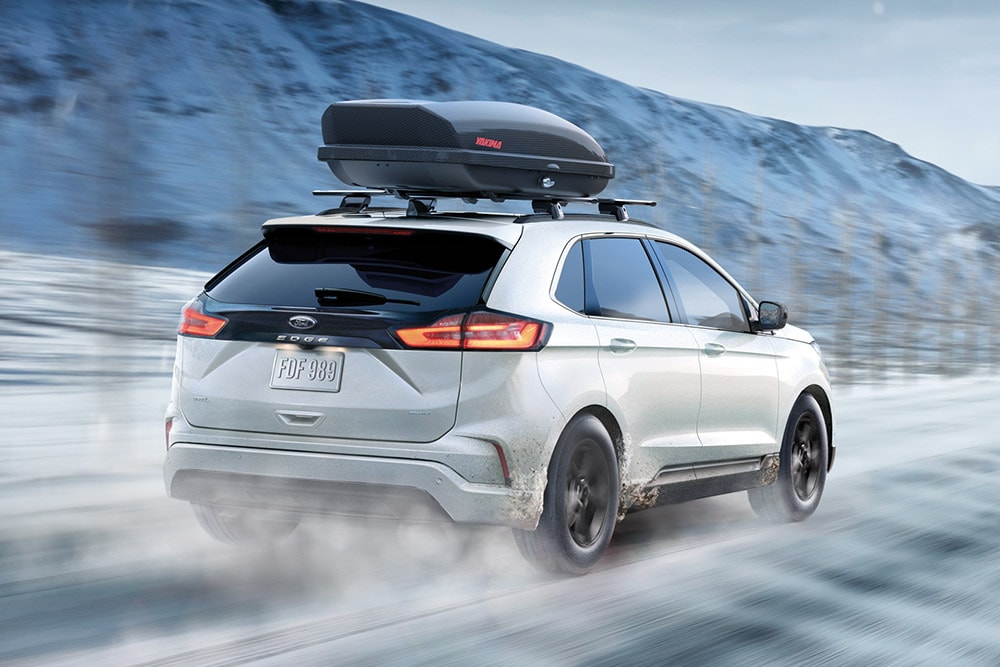 Bílý Ford Edge s boxem na střeše jede po zasněžené horské silnici, rozmazaný pohyb.