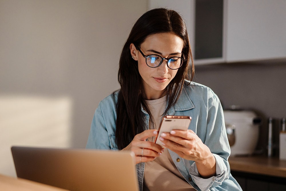 Osoba v brýlích se dívá na smartphone, s notebookem na dřevěném stole.