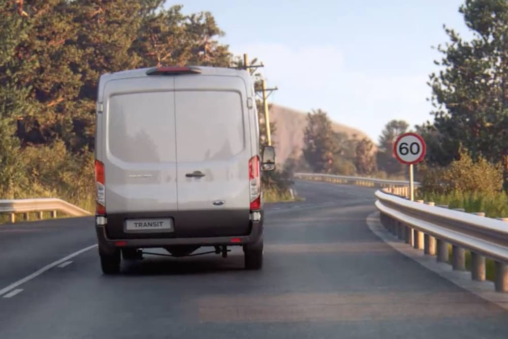 Dodávka Ford Transit jede po silnici kolem dopravní značky, při pohledu zezadu.